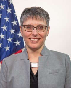 Headshot of Laura Stanton in front of an American flag. She is wearing black glasses, a blazer, and a smile. 